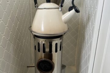 A white, vintage-style kettle with a black handle and spout is placed on top of a metallic stove with ventilation slots. The stove is positioned in the corner of a room with white patterned tile walls. A metal chimney pipe extends from the back of the stove upwards.