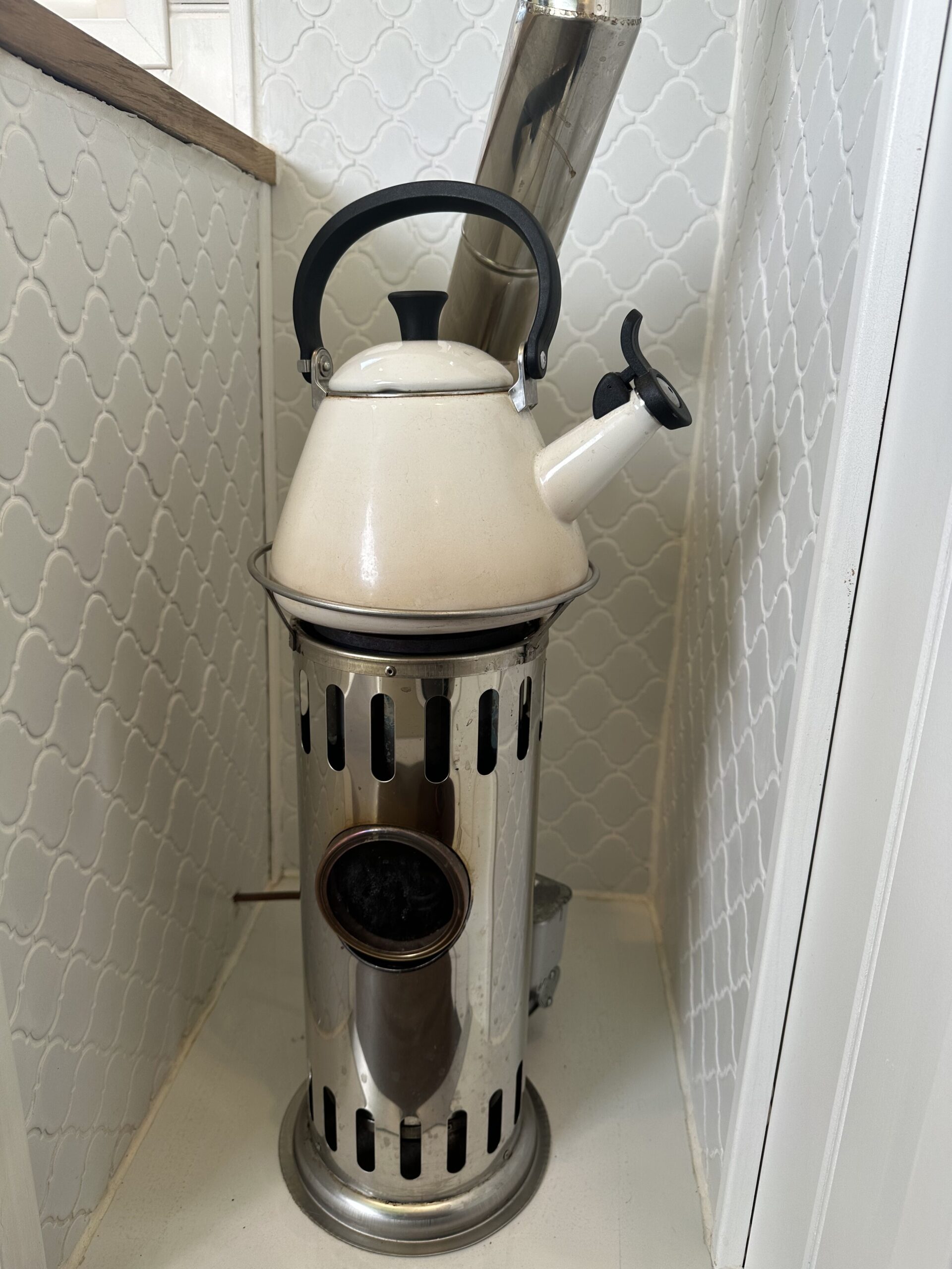 A white, vintage-style kettle with a black handle and spout is placed on top of a metallic stove with ventilation slots. The stove is positioned in the corner of a room with white patterned tile walls. A metal chimney pipe extends from the back of the stove upwards.
