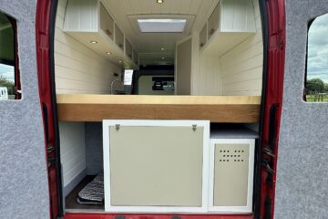 A red van with its rear doors open, revealing a built-in kitchen. The interior features white cabinetry, a wooden countertop, and a light fixture overhead. The floor is finished in darker wood. The sky appears cloudy, and green grass surrounds the van.
