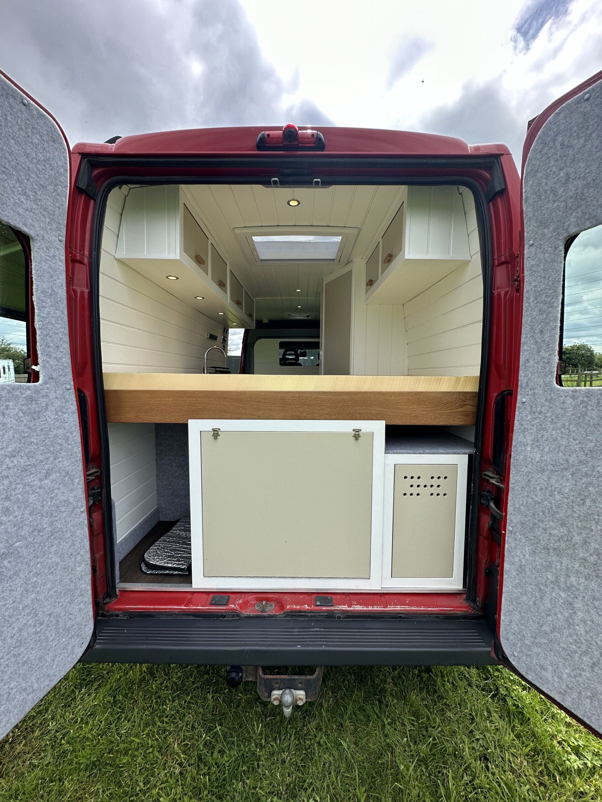 A red van with its rear doors open, revealing a built-in kitchen. The interior features white cabinetry, a wooden countertop, and a light fixture overhead. The floor is finished in darker wood. The sky appears cloudy, and green grass surrounds the van.