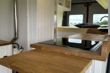 A cozy, modern RV kitchen with light wood countertops and white paneled walls. It features a black stovetop embedded in the counter, a pull-out wooden extension for extra workspace, and a chimney stove. Shelves and storage compartments are built into the walls, with a large window in the background.