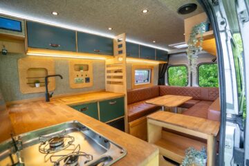 Interior view of a modern camper van with a compact yet stylish setup. The left side features a kitchen area with a stainless steel sink and stove on wooden countertops. The right side has a cozy seating area with a table and cushioned benches, and the space is well-lit with overhead lights.