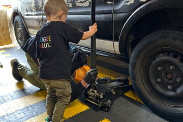 A young child stands next to a black vehicle holding a car jack handle, assisting an adult who is lying on their back underneath the car. The child wears a black shirt with colorful text, and the scene takes place on a black-and-yellow checkered floor in a garage.