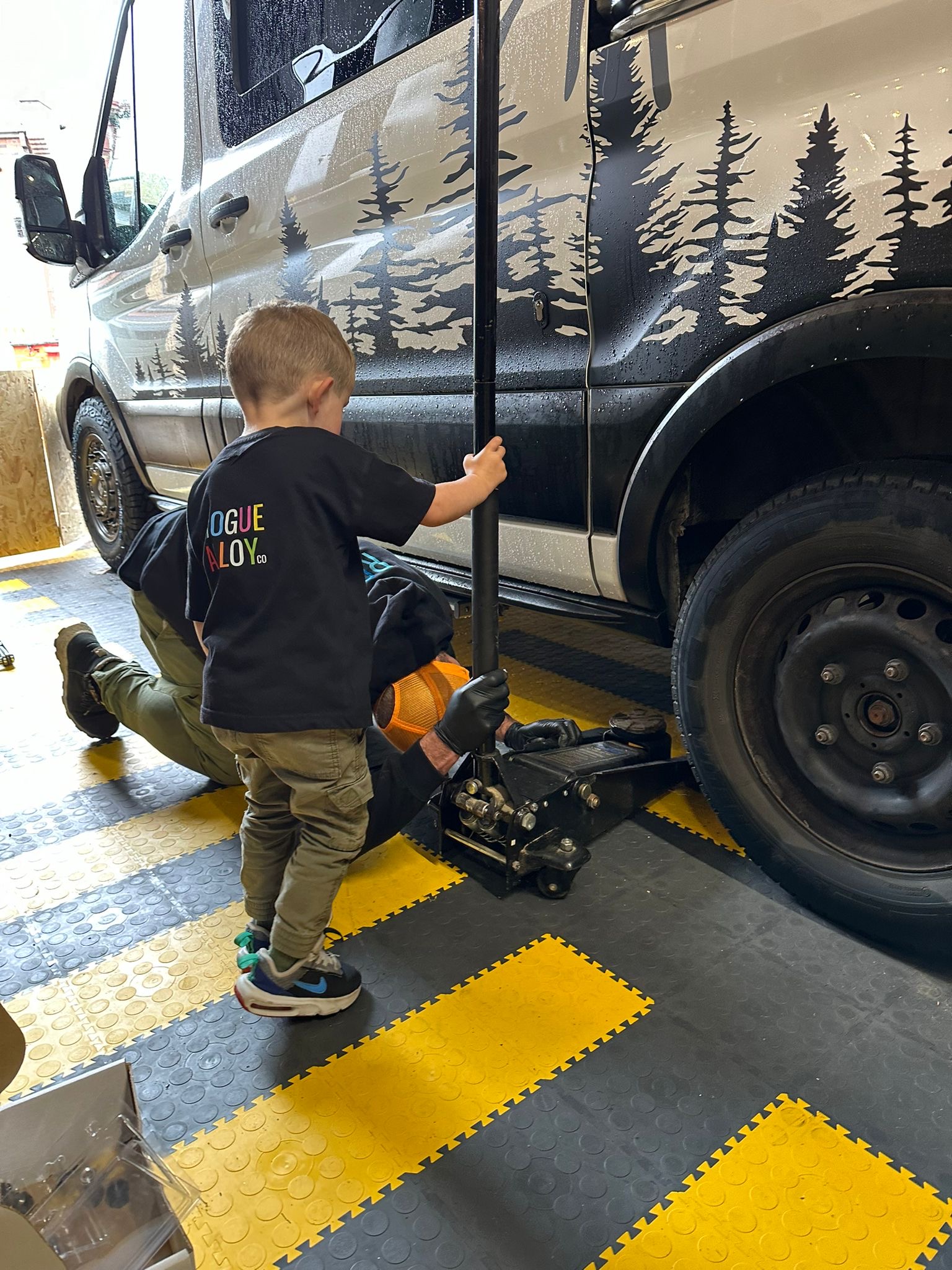 A young child stands next to a black vehicle holding a car jack handle, assisting an adult who is lying on their back underneath the car. The child wears a black shirt with colorful text, and the scene takes place on a black-and-yellow checkered floor in a garage.