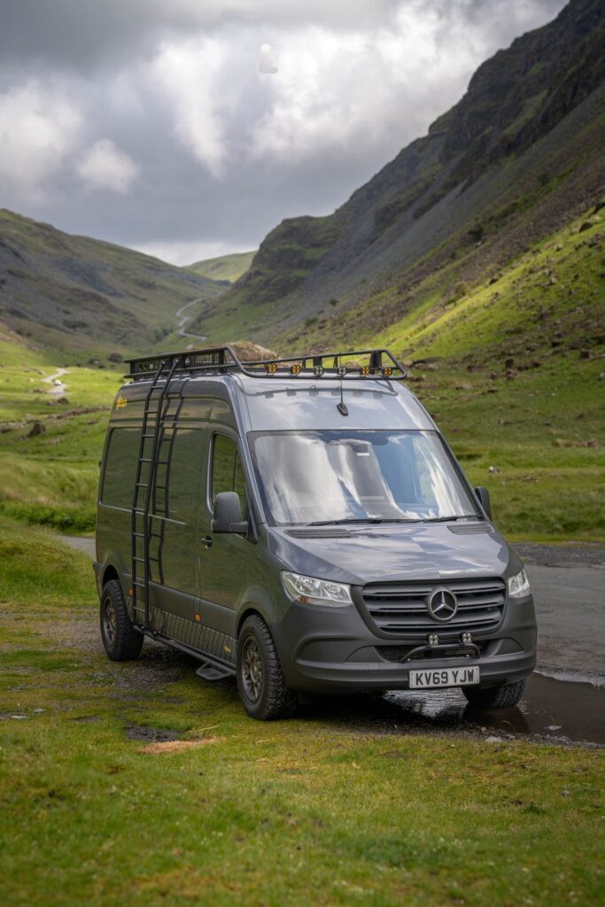 A gray Mercedes-Benz Sprinter van is parked on a grassy area near a winding road in a picturesque, mountainous landscape. The van has a roof rack equipped with storage and a ladder on its side. The sky is cloudy, casting a dramatic backdrop over the green hills and rugged terrain.