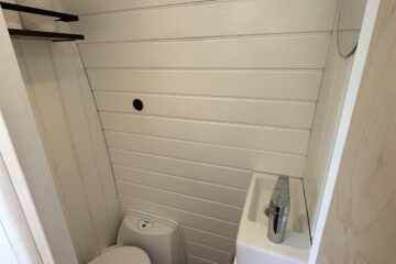 A compact bathroom features a white composting toilet in the corner next to a small white sink with a modern silver faucet. The walls are clad in white horizontal wood paneling. A small round mirror is mounted above the sink, and a black hook is installed on the wall opposite the toilet.