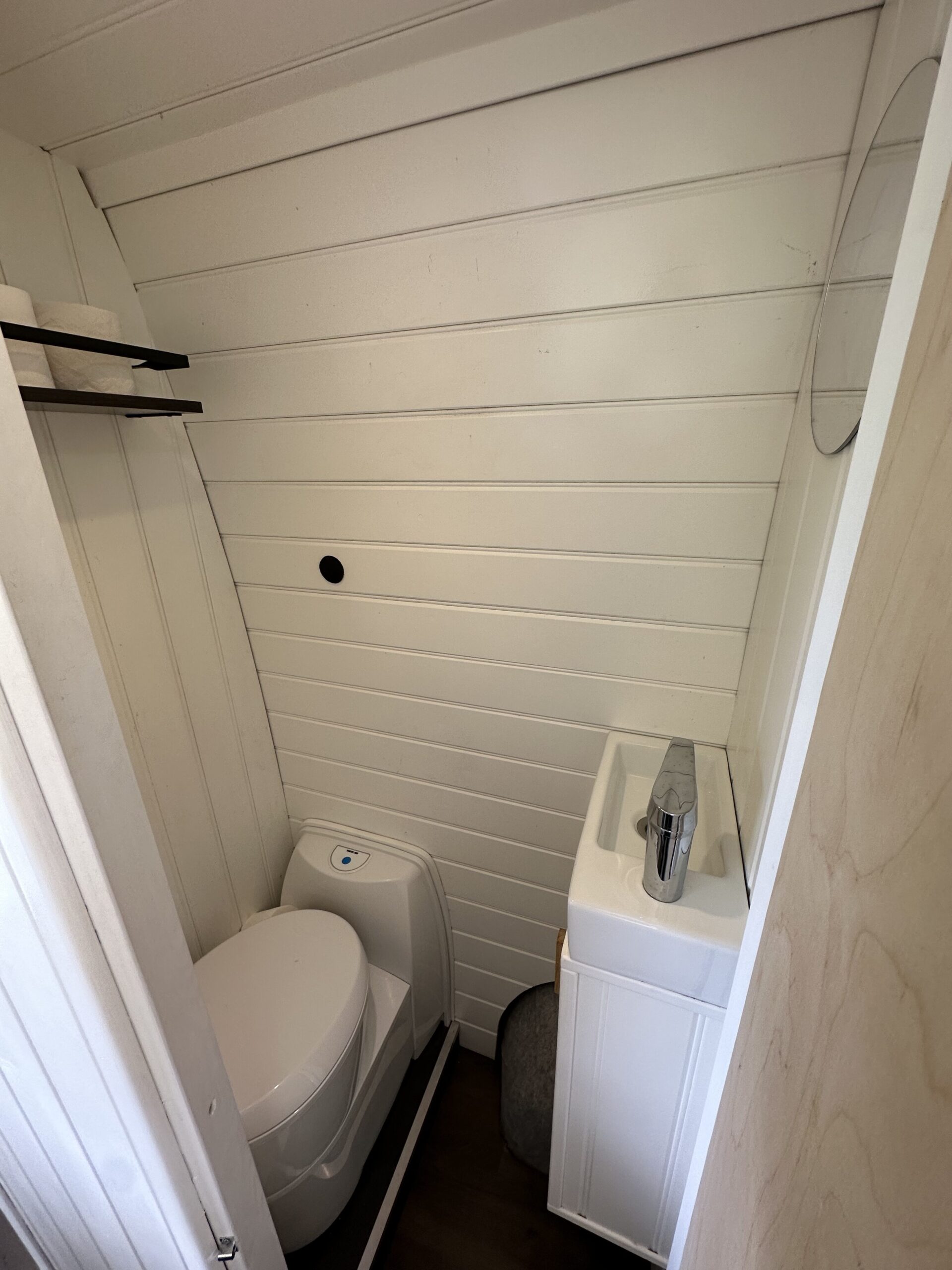 A compact bathroom features a white composting toilet in the corner next to a small white sink with a modern silver faucet. The walls are clad in white horizontal wood paneling. A small round mirror is mounted above the sink, and a black hook is installed on the wall opposite the toilet.
