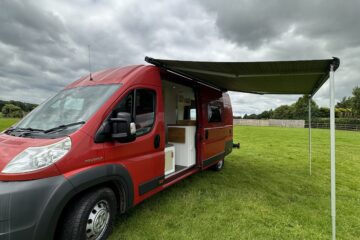 A bright red camper van with an extended awning is parked on lush green grass under a partly cloudy sky. The van's side door is open, revealing a white interior with a kitchen counter. In the background, there is a wooden fence and trees along the horizon.