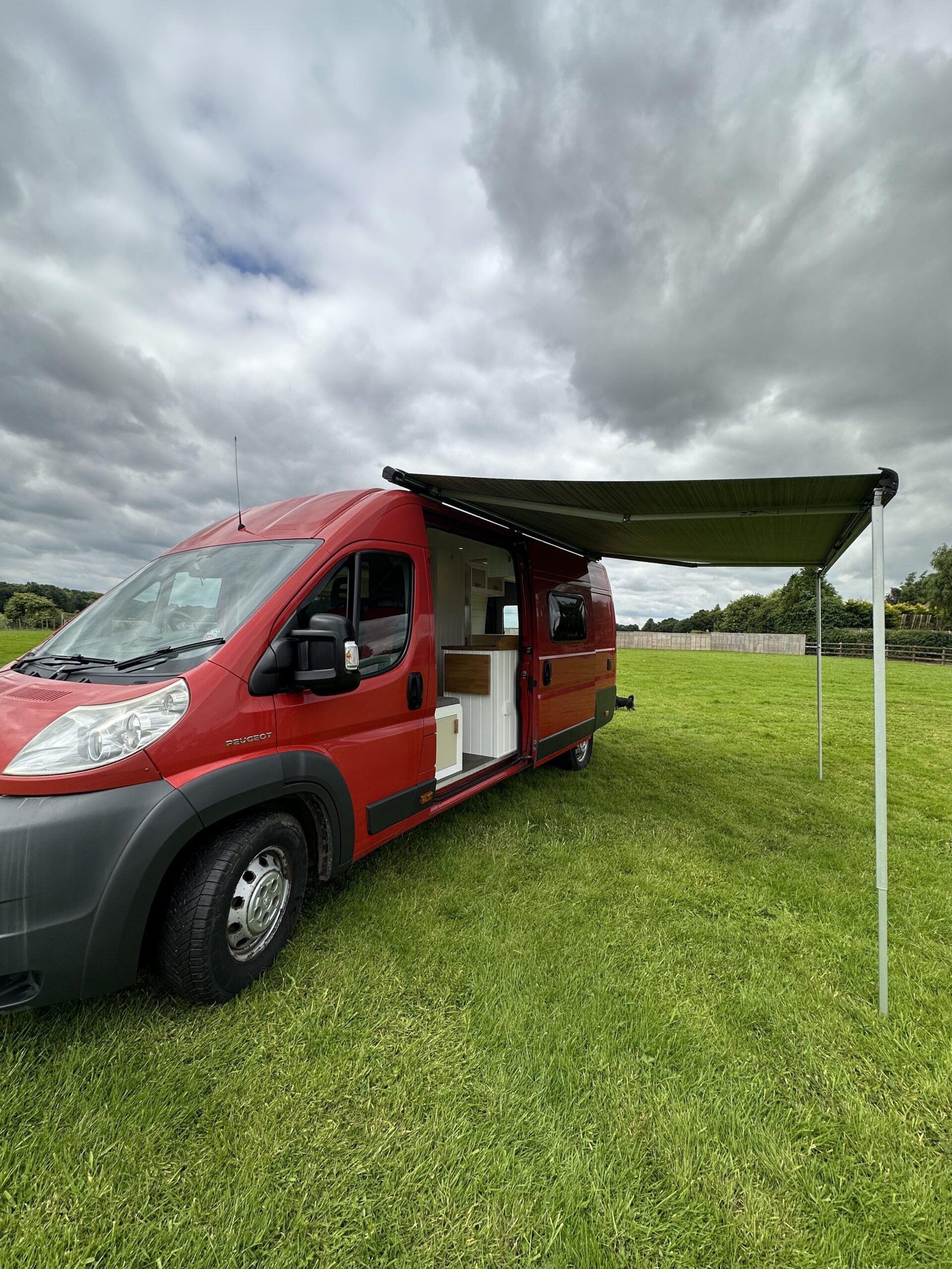 A bright red camper van with an extended awning is parked on lush green grass under a partly cloudy sky. The van's side door is open, revealing a white interior with a kitchen counter. In the background, there is a wooden fence and trees along the horizon.