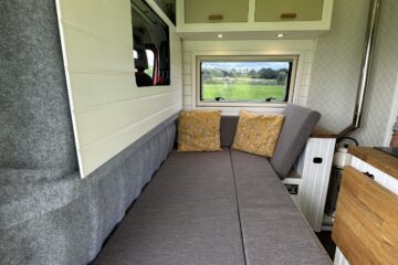 Interior of a camper van featuring a grey cushioned bench with yellow patterned pillows against the backrest, convertible into a bed. Above is a white cabinet with two doors, and the ceiling has a round vent. A window reveals a lush green field outside, and wood accents add warmth.