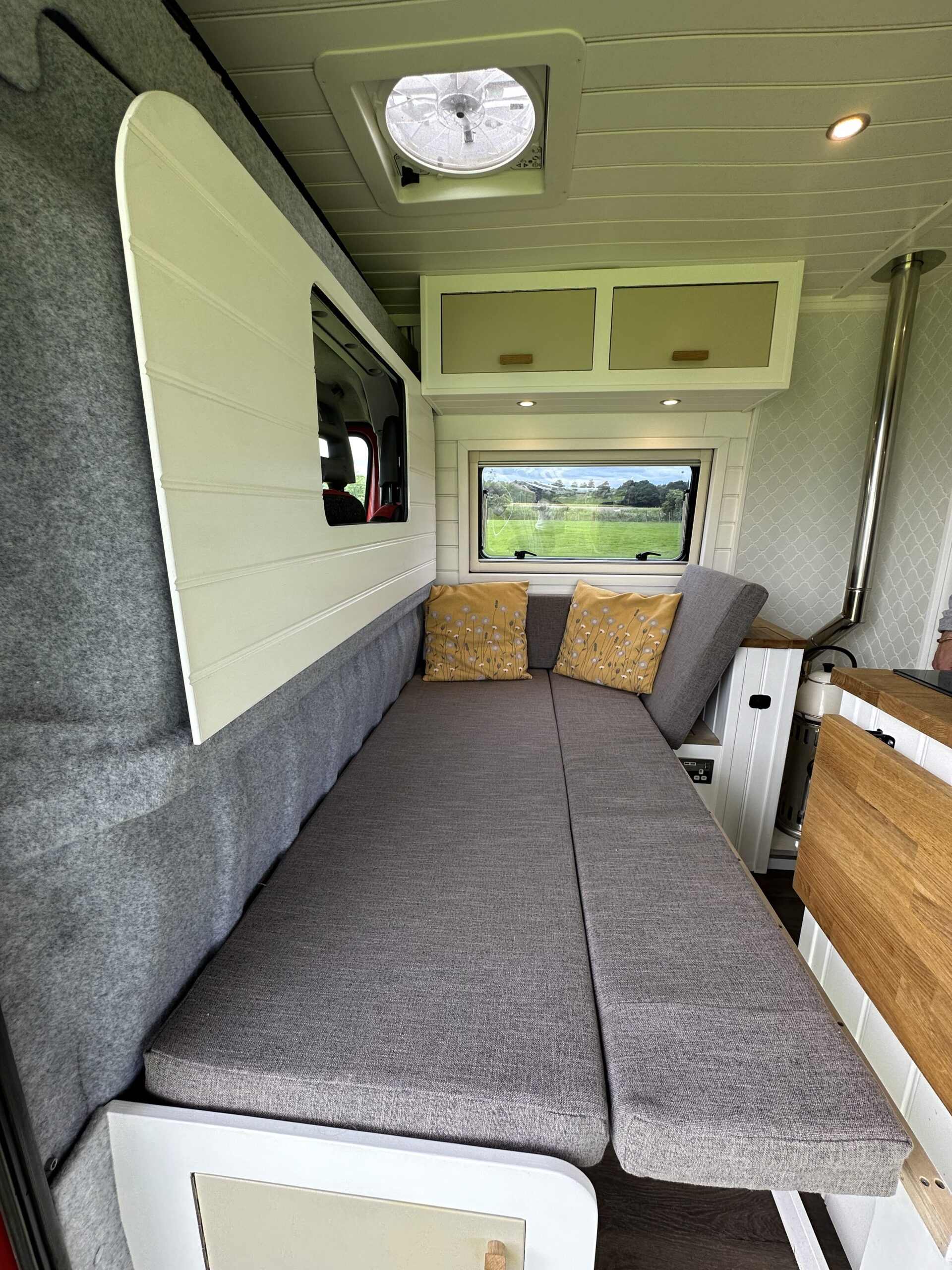 Interior of a camper van featuring a grey cushioned bench with yellow patterned pillows against the backrest, convertible into a bed. Above is a white cabinet with two doors, and the ceiling has a round vent. A window reveals a lush green field outside, and wood accents add warmth.