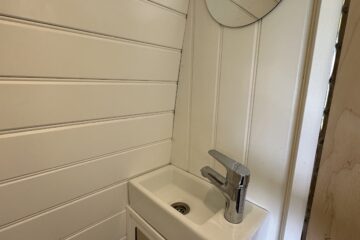 A small, white bathroom sink is positioned in the corner of a room with white wood-paneled walls. Above the sink is a round mirror. The sink features a modern, chrome faucet and has a small cabinet beneath it with a light-colored door and wooden handle.