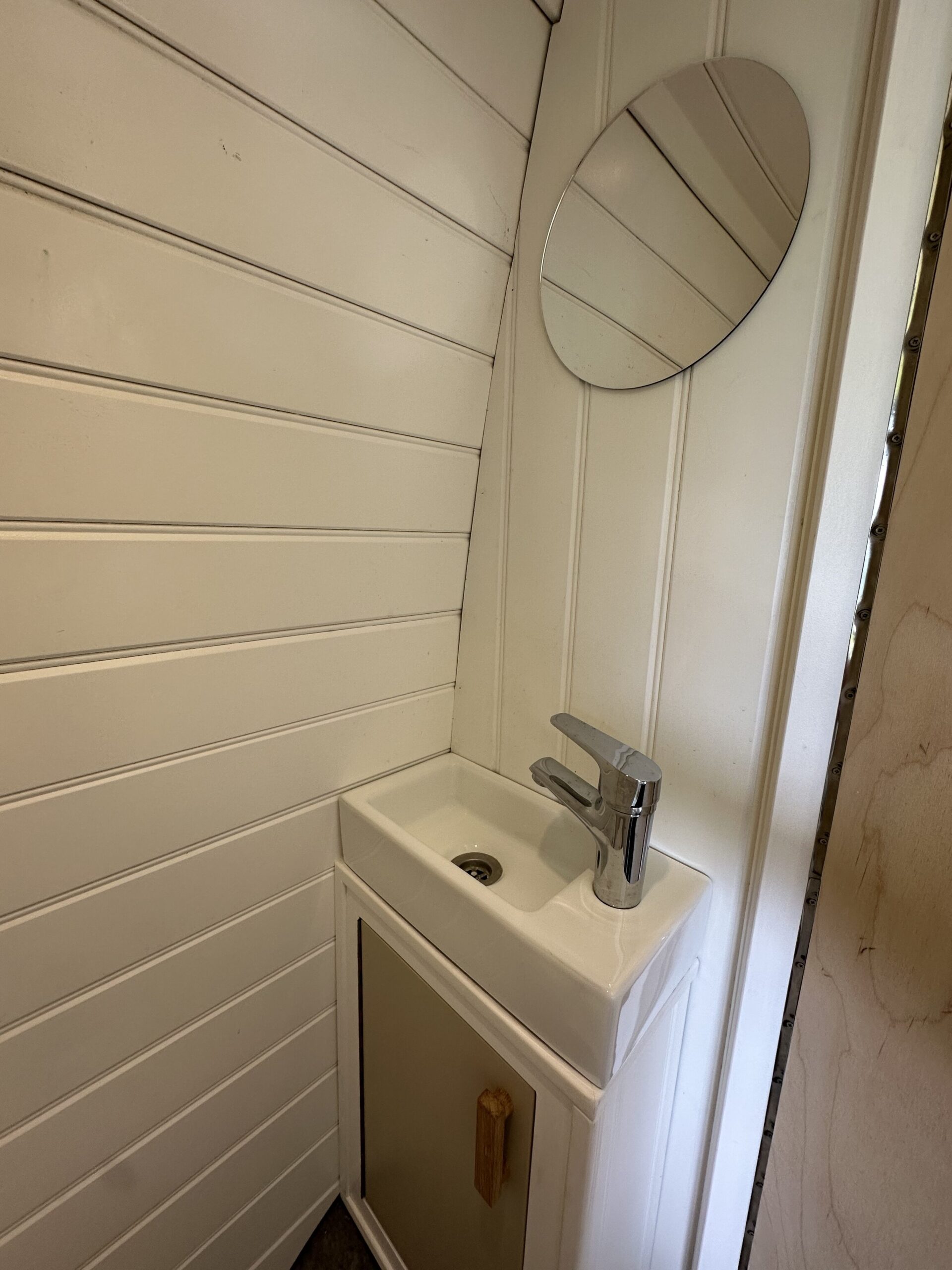 A small, white bathroom sink is positioned in the corner of a room with white wood-paneled walls. Above the sink is a round mirror. The sink features a modern, chrome faucet and has a small cabinet beneath it with a light-colored door and wooden handle.