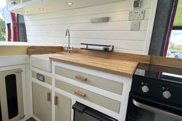 A modern campervan kitchen features white cabinetry with wooden handles and a wood countertop. It has multiple drawers and cabinets, a small fridge, an oven with a stove, an overhead storage area, and a silver faucet with a sink. The walls are light-toned with shiplap paneling.