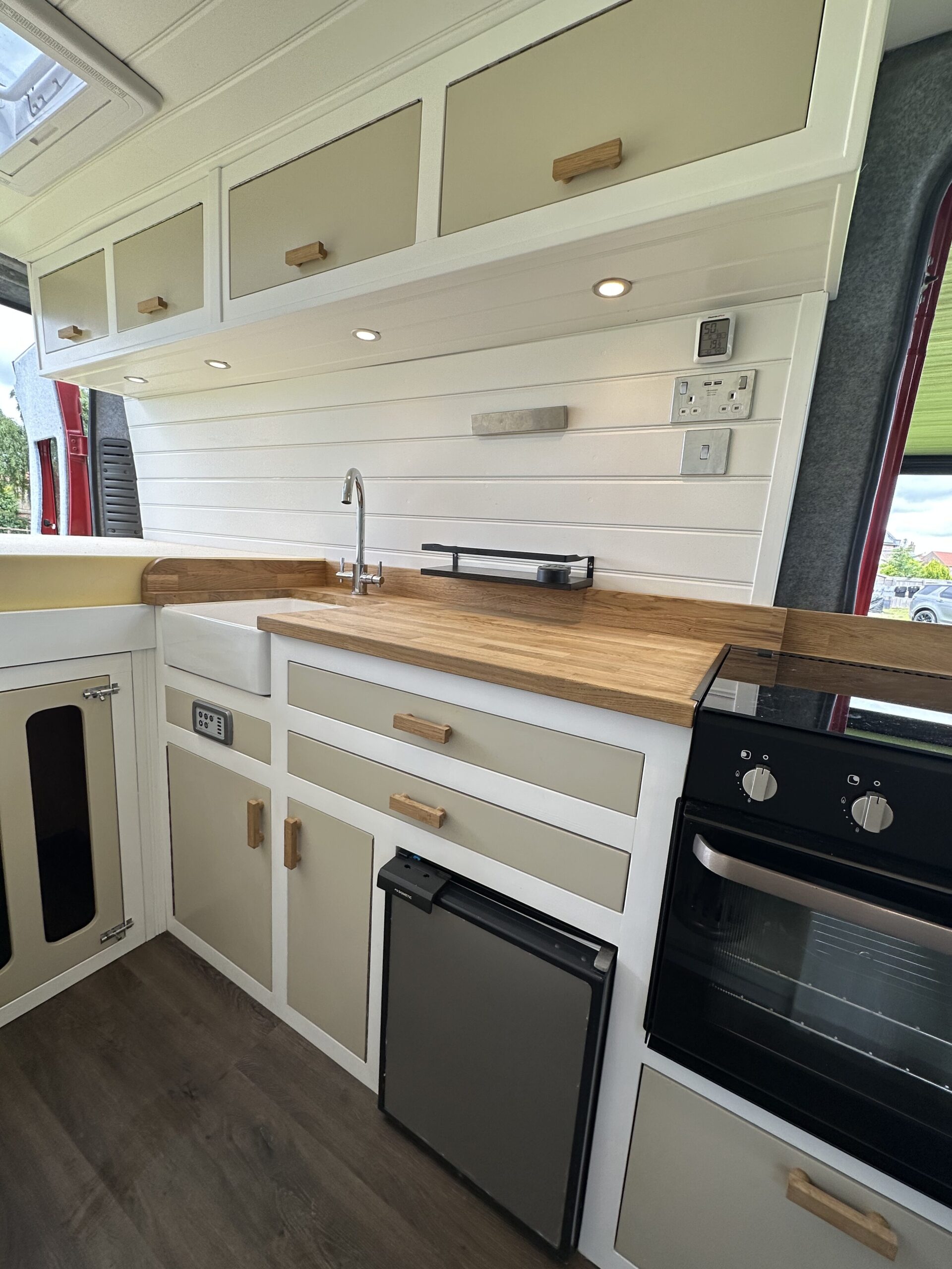 A modern campervan kitchen features white cabinetry with wooden handles and a wood countertop. It has multiple drawers and cabinets, a small fridge, an oven with a stove, an overhead storage area, and a silver faucet with a sink. The walls are light-toned with shiplap paneling.
