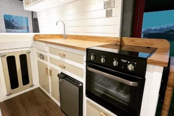 A modern, compact kitchen inside a vehicle, featuring white cabinets with light wood trim. The setup includes a stainless steel sink with a wooden countertop, an under-counter fridge, a small oven, and overhead storage cabinets. Recessed lighting under the cabinets illuminates the area. The floor has a wood finish.