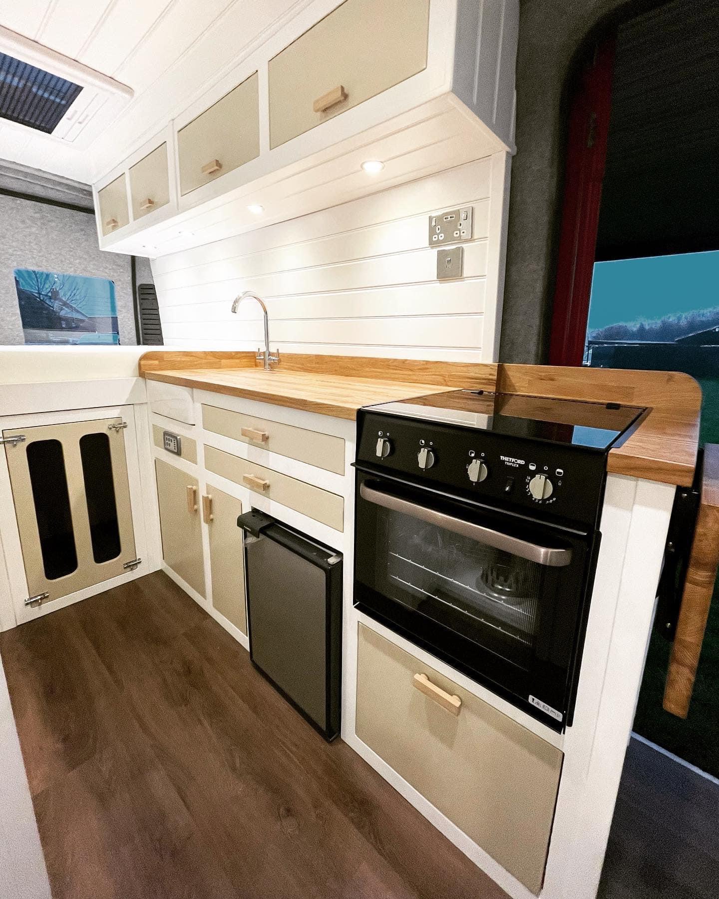 A modern, compact kitchen inside a vehicle, featuring white cabinets with light wood trim. The setup includes a stainless steel sink with a wooden countertop, an under-counter fridge, a small oven, and overhead storage cabinets. Recessed lighting under the cabinets illuminates the area. The floor has a wood finish.