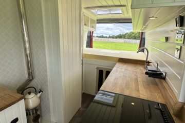 A compact, modern camper van kitchen featuring wood countertops, a stove, sink, and small oven. There is a cylindrical wood-burning stove with a chimney. The interior is primarily white, with wood elements and a diamond-pattern backsplash. Sunlight illuminates the room through windows.