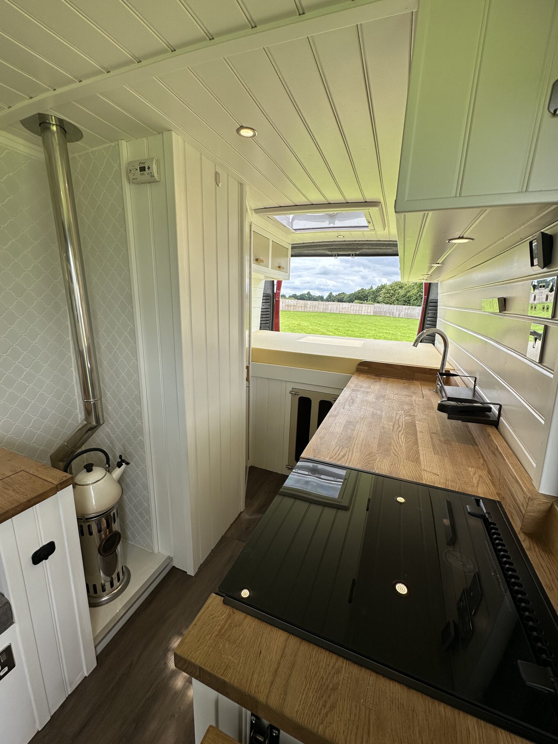 A compact, modern camper van kitchen featuring wood countertops, a stove, sink, and small oven. There is a cylindrical wood-burning stove with a chimney. The interior is primarily white, with wood elements and a diamond-pattern backsplash. Sunlight illuminates the room through windows.