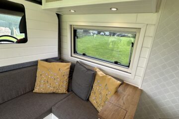 The image shows the interior of a camper van with a cozy corner seating area. The seating has grey cushions and is accented with yellow pillows featuring a floral design. Above the seating, there's a window with a view of a grassy field and overhead cabinets. A small counter and a kettle are visible to the right.