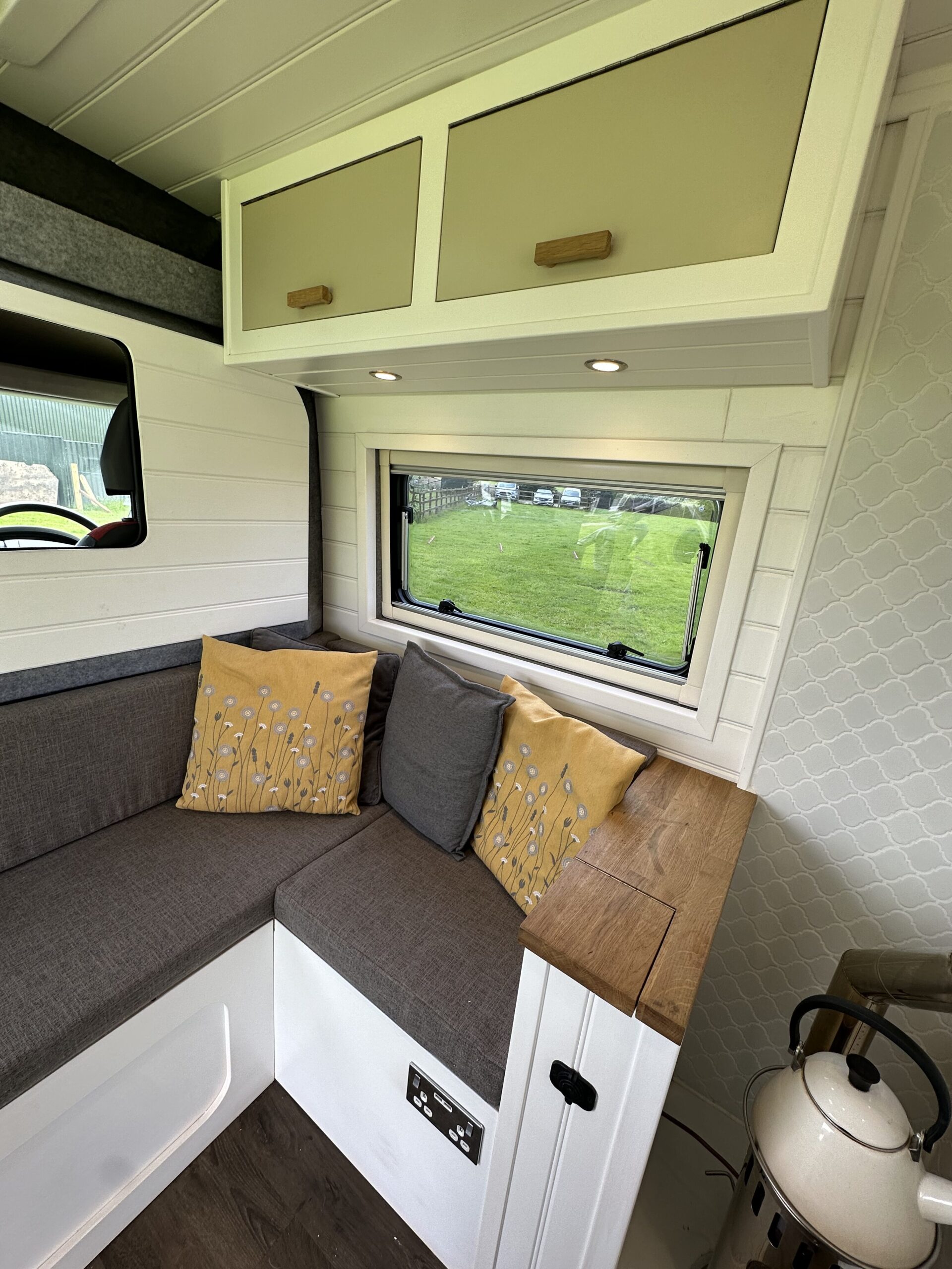 The image shows the interior of a camper van with a cozy corner seating area. The seating has grey cushions and is accented with yellow pillows featuring a floral design. Above the seating, there's a window with a view of a grassy field and overhead cabinets. A small counter and a kettle are visible to the right.