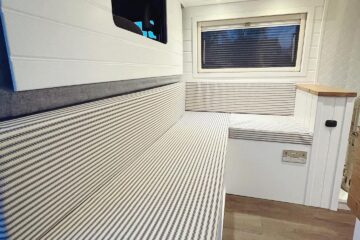 Interior of a modern camper van with a cozy seating area. The seats have striped upholstery and are built along the length of the van. There's a window with a blind above the seat, and overhead storage cabinets with cream-colored doors and subtle downlights. The floor is a light wood finish.