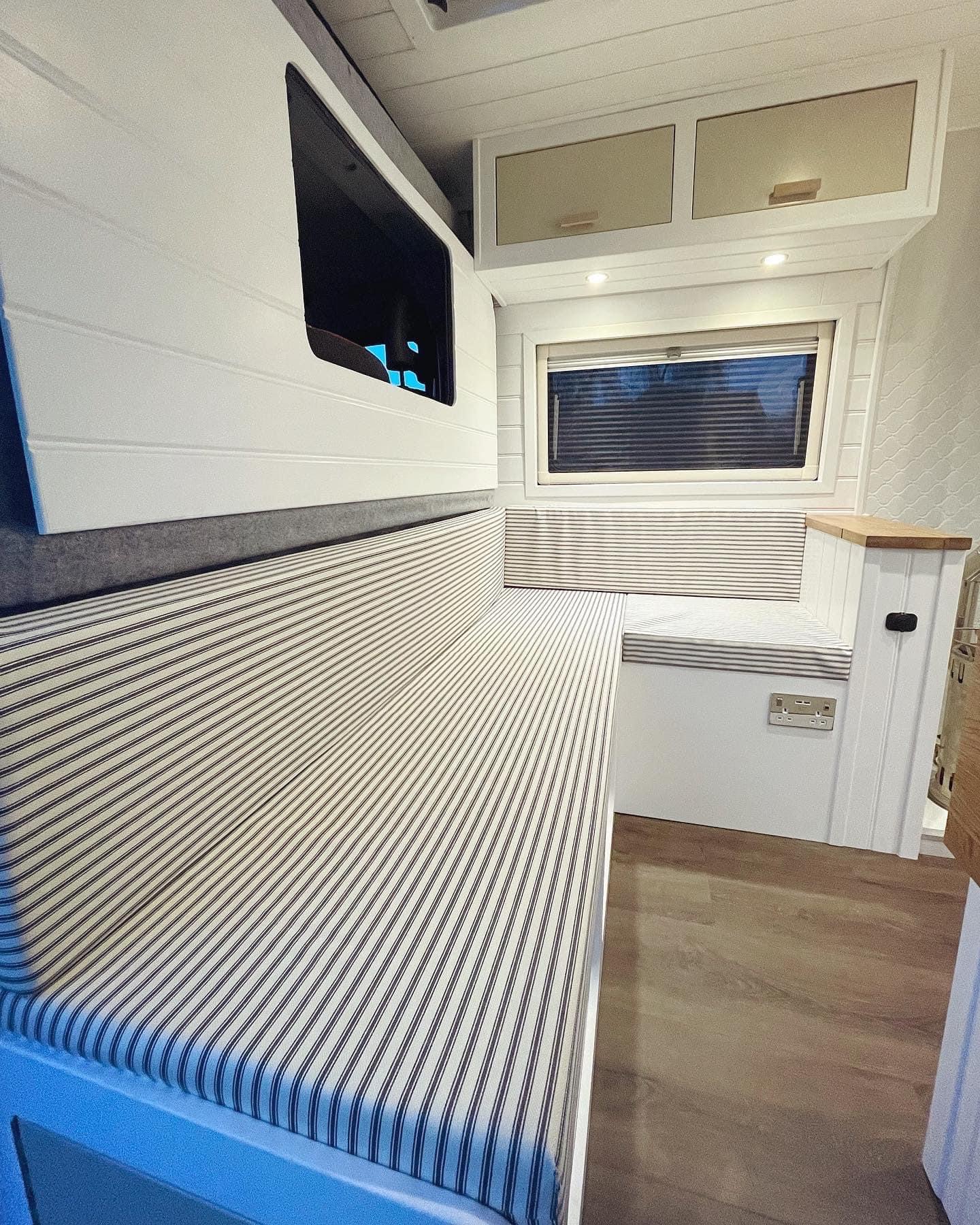 Interior of a modern camper van with a cozy seating area. The seats have striped upholstery and are built along the length of the van. There's a window with a blind above the seat, and overhead storage cabinets with cream-colored doors and subtle downlights. The floor is a light wood finish.