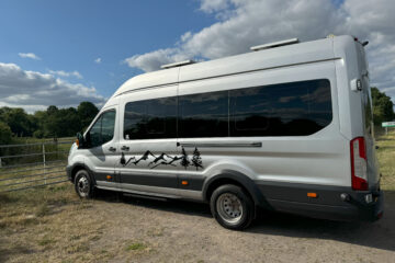 A white camper van is parked near a metal gate on a grassy area. The van features black mountain and tree decals along its side. The sky above is partly cloudy, casting soft shadows on the vehicle. The background includes trees and distant greenery under a blue sky.