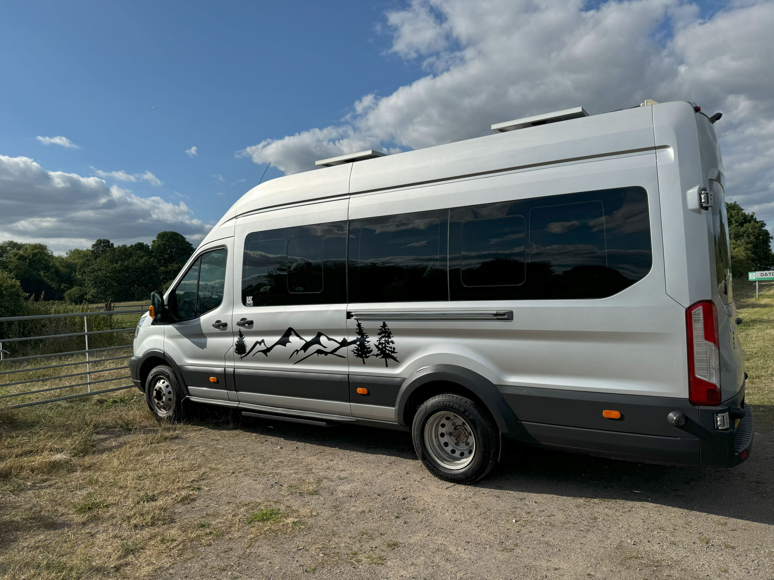 A white camper van is parked near a metal gate on a grassy area. The van features black mountain and tree decals along its side. The sky above is partly cloudy, casting soft shadows on the vehicle. The background includes trees and distant greenery under a blue sky.