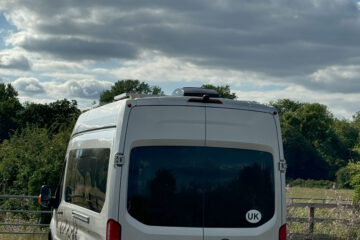 A white Ford Transit van with a UK sticker is parked in a rural area next to a wooden fence. The van has a license plate reading "NE55 VAN." In the background, lush green trees and bushes are visible under a partly cloudy sky. Graffiti is seen on the fence.