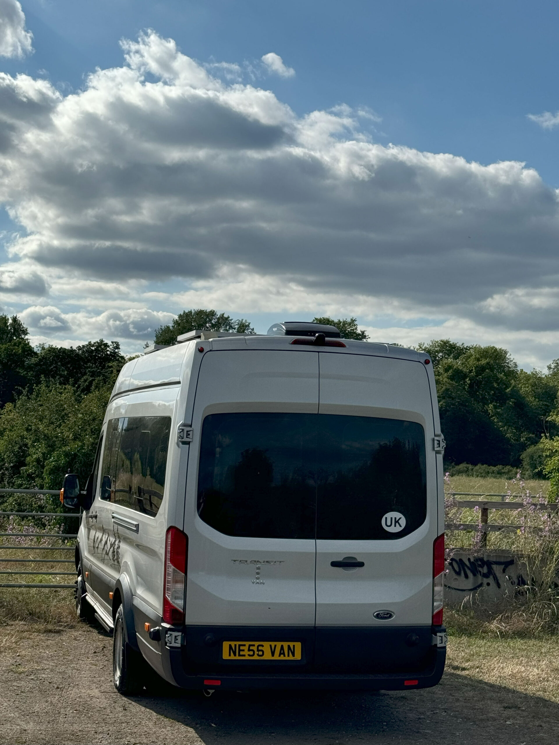 A white Ford Transit van with a UK sticker is parked in a rural area next to a wooden fence. The van has a license plate reading 