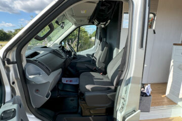 A silver van with its driver-side door open, showing the front interior. The seats are black, and a black bag hangs behind the driver's seat. A white cabinet is visible inside the van, indicating its use for camping or travel. The outside landscape features green shrubs and a bright blue sky.