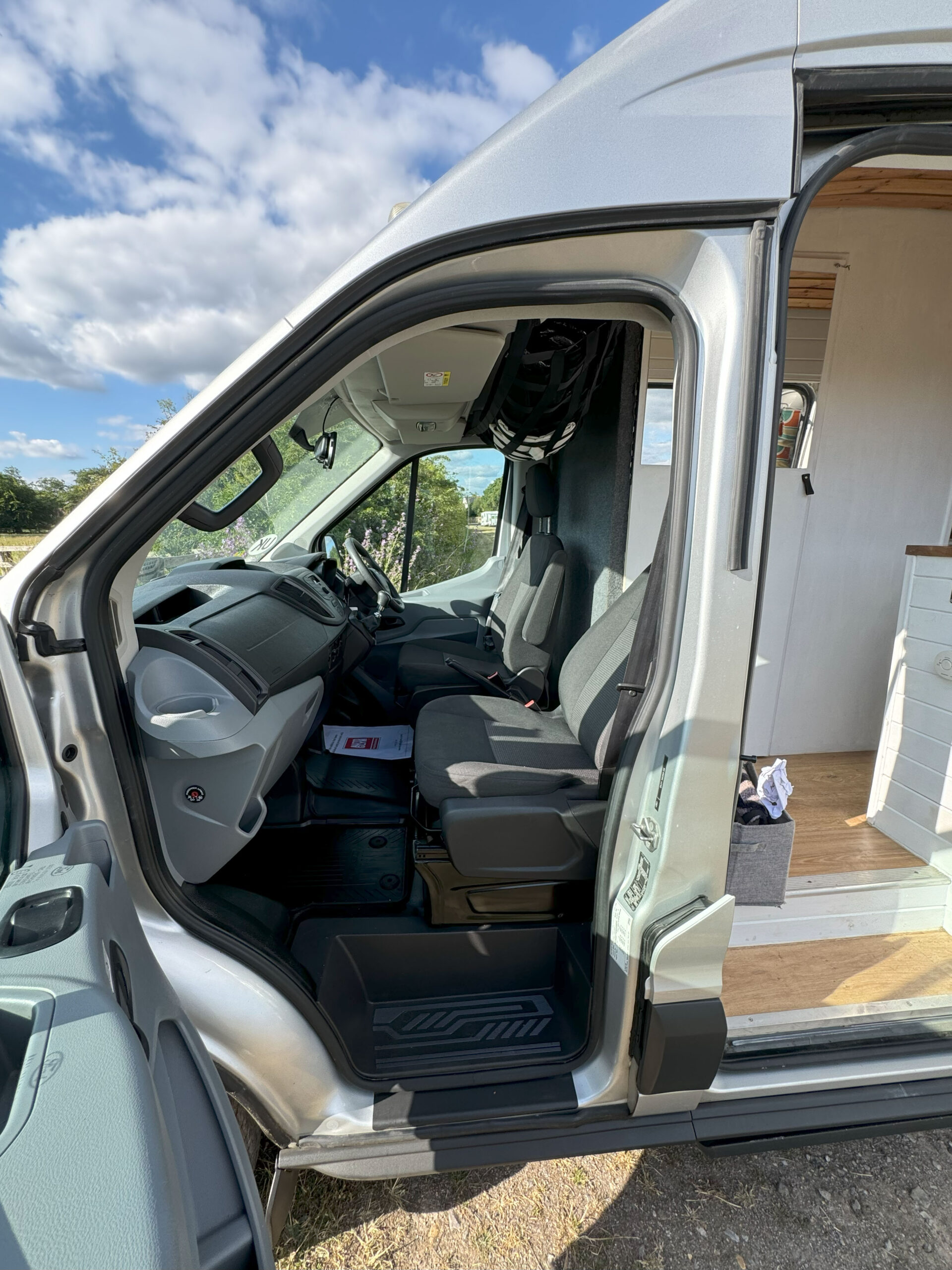 A silver van with its driver-side door open, showing the front interior. The seats are black, and a black bag hangs behind the driver's seat. A white cabinet is visible inside the van, indicating its use for camping or travel. The outside landscape features green shrubs and a bright blue sky.