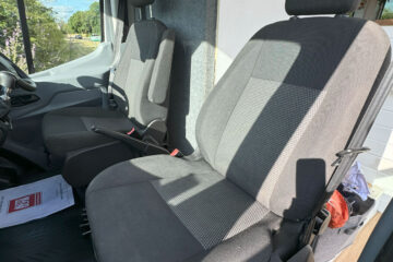 Interior of a vehicle showing two front seats. The seats are upholstered in dark fabric with lighter mesh-like inserts in the center. The driver's seat has adjustable armrests. The sun casts shadows through the windows. Some papers are seen on the floor near the driver's seat.