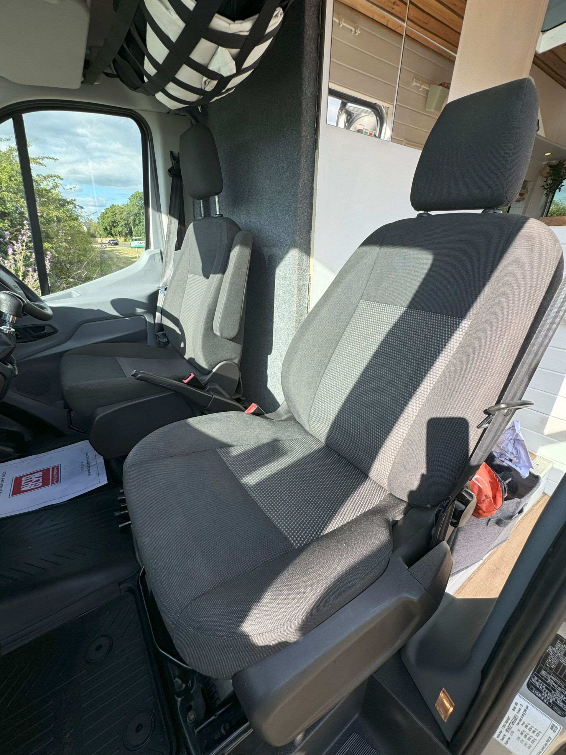 Interior of a vehicle showing two front seats. The seats are upholstered in dark fabric with lighter mesh-like inserts in the center. The driver's seat has adjustable armrests. The sun casts shadows through the windows. Some papers are seen on the floor near the driver's seat.