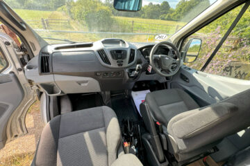 Color photograph of a van's interior taken from the rear. It shows two gray front seats, a dashboard with a steering wheel, and various controls. Out the windshield, a scenic view of a lush green rural landscape is visible, with trees, a wooden fence, and blue sky.