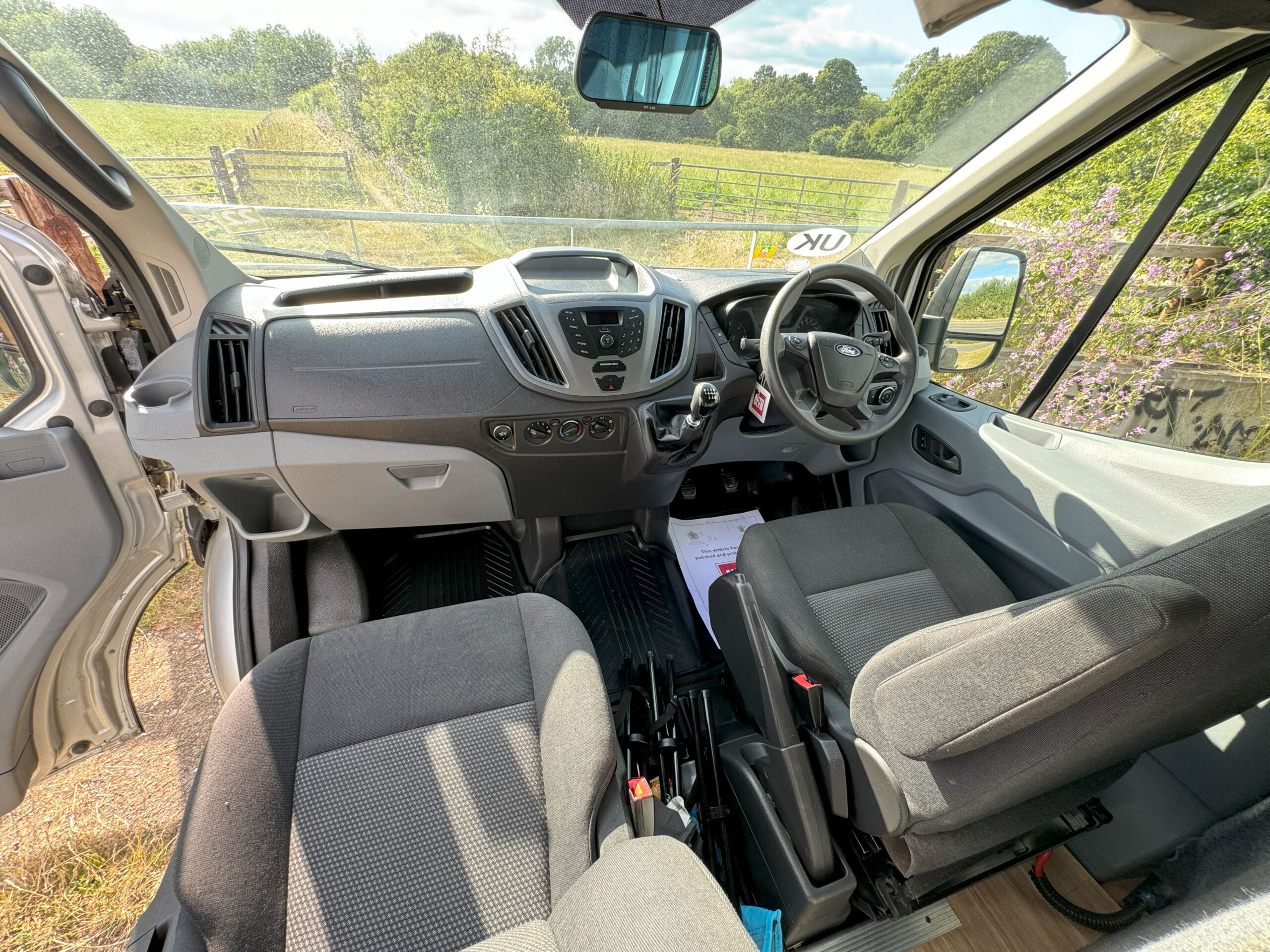 Color photograph of a van's interior taken from the rear. It shows two gray front seats, a dashboard with a steering wheel, and various controls. Out the windshield, a scenic view of a lush green rural landscape is visible, with trees, a wooden fence, and blue sky.