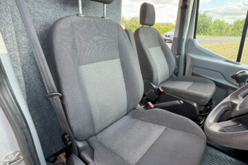 Interior view of a vehicle's front cab, featuring two gray fabric seats with headrests. The driver's seat is on the right, with the steering wheel partially visible. The passenger seat is on the left. The floor has black mats, and a paper with red text is on the floor near the dashboard.