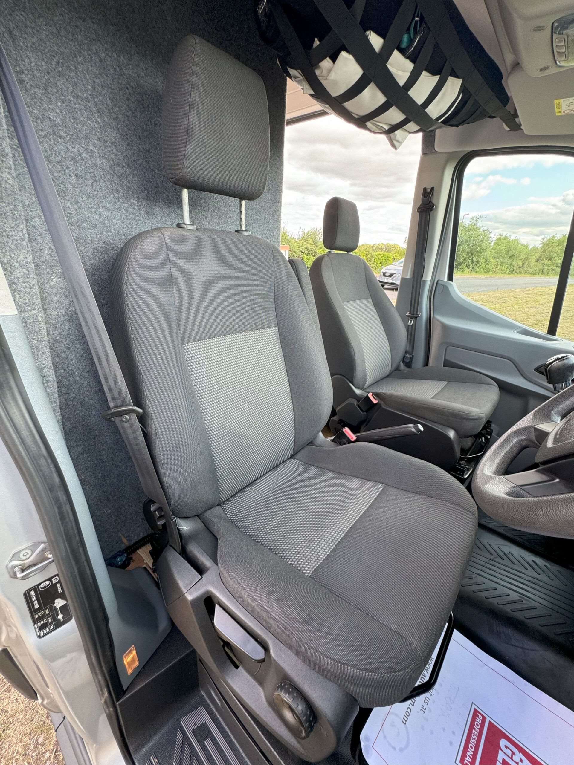 Interior view of a vehicle's front cab, featuring two gray fabric seats with headrests. The driver's seat is on the right, with the steering wheel partially visible. The passenger seat is on the left. The floor has black mats, and a paper with red text is on the floor near the dashboard.