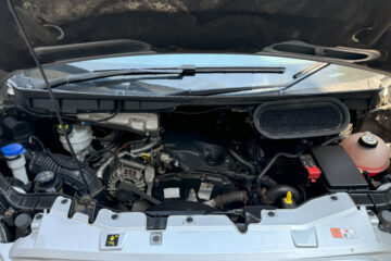 Close-up image of a car engine compartment with the hood open. Various engine components are visible, including the battery, coolant reservoir, air intake, and other mechanical parts. The engine is clean, and the arrangement of parts displays typical modern vehicle engineering design.