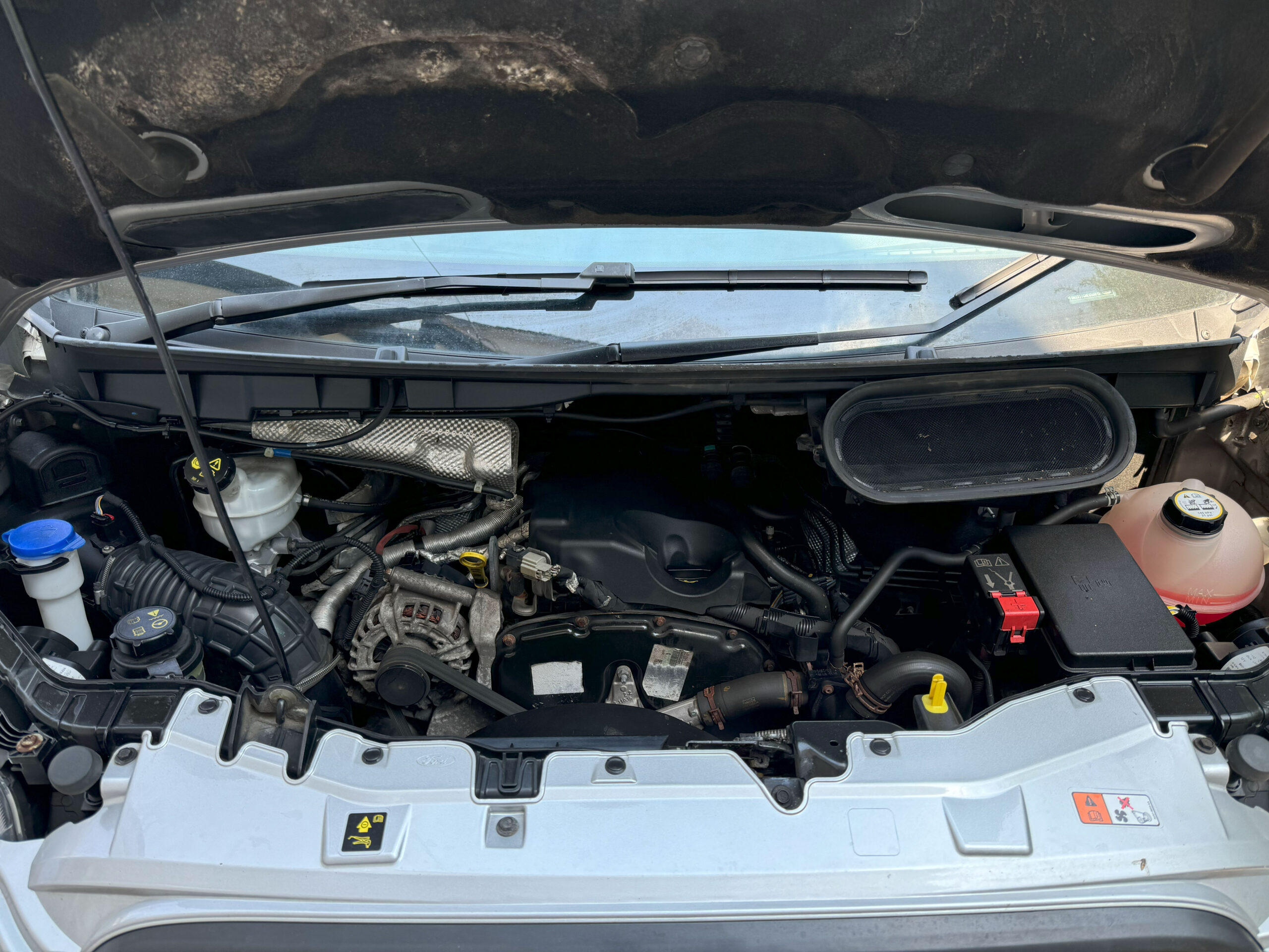 Close-up image of a car engine compartment with the hood open. Various engine components are visible, including the battery, coolant reservoir, air intake, and other mechanical parts. The engine is clean, and the arrangement of parts displays typical modern vehicle engineering design.