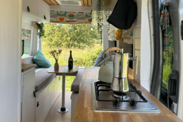 Interior of a converted camper van with wood-paneled ceiling, a skylight, and white walls. The kitchen area has a stove on a wooden countertop and plants hanging near the window. A wine bottle and glasses are on a small table. The back area features seats with colorful cushions and a large window.