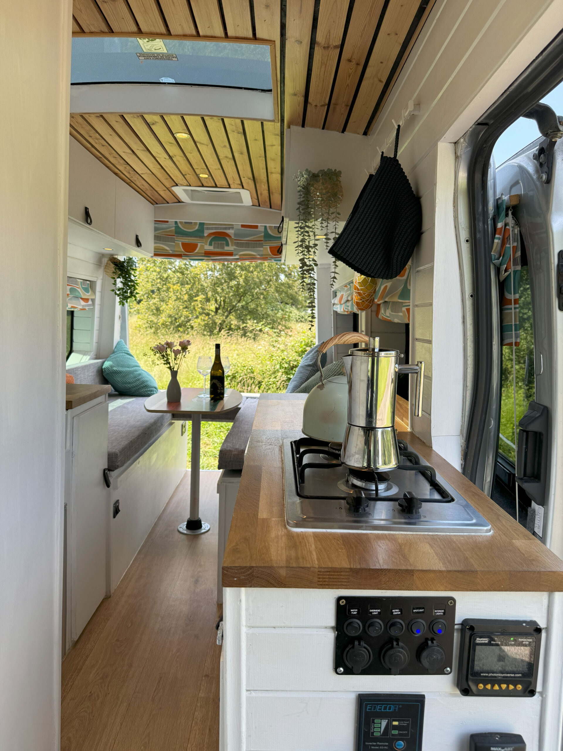 Interior of a converted camper van with wood-paneled ceiling, a skylight, and white walls. The kitchen area has a stove on a wooden countertop and plants hanging near the window. A wine bottle and glasses are on a small table. The back area features seats with colorful cushions and a large window.