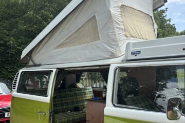 A green and white vintage camper van is parked outdoors. The pop-up roof is extended with canvas siding and a mesh window. The side door is open, revealing a cozy interior with plaid upholstery on the seats. Trees and another red vehicle are visible in the background.