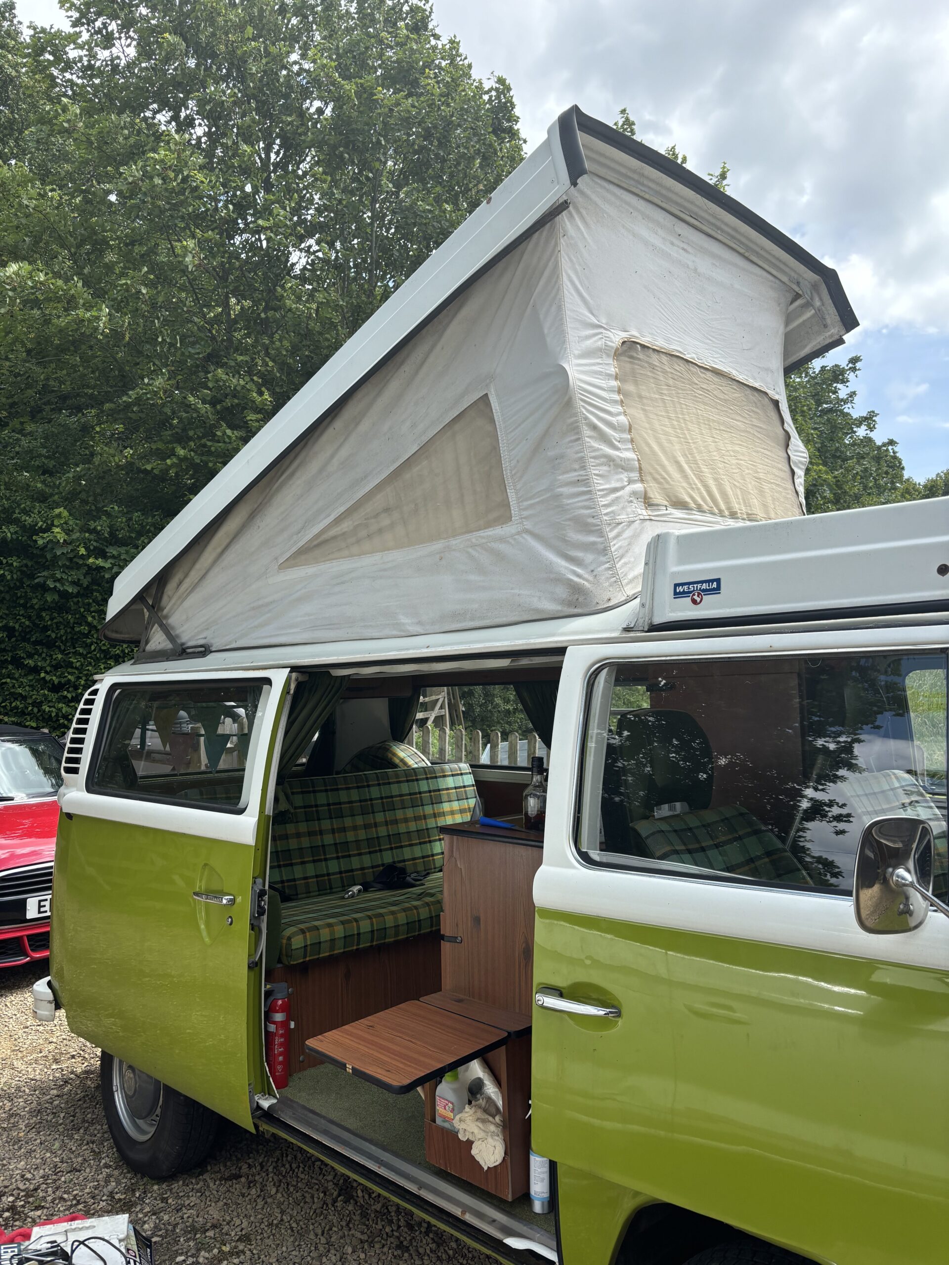 A green and white vintage camper van is parked outdoors. The pop-up roof is extended with canvas siding and a mesh window. The side door is open, revealing a cozy interior with plaid upholstery on the seats. Trees and another red vehicle are visible in the background.