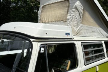 A classic green and white Volkswagen camper van with a raised pop-up roof is parked outdoors. The pop-up roof has a beige canvas with a window, allowing for additional headroom inside. The side windows are slightly open, and the surrounding area features lush green trees.