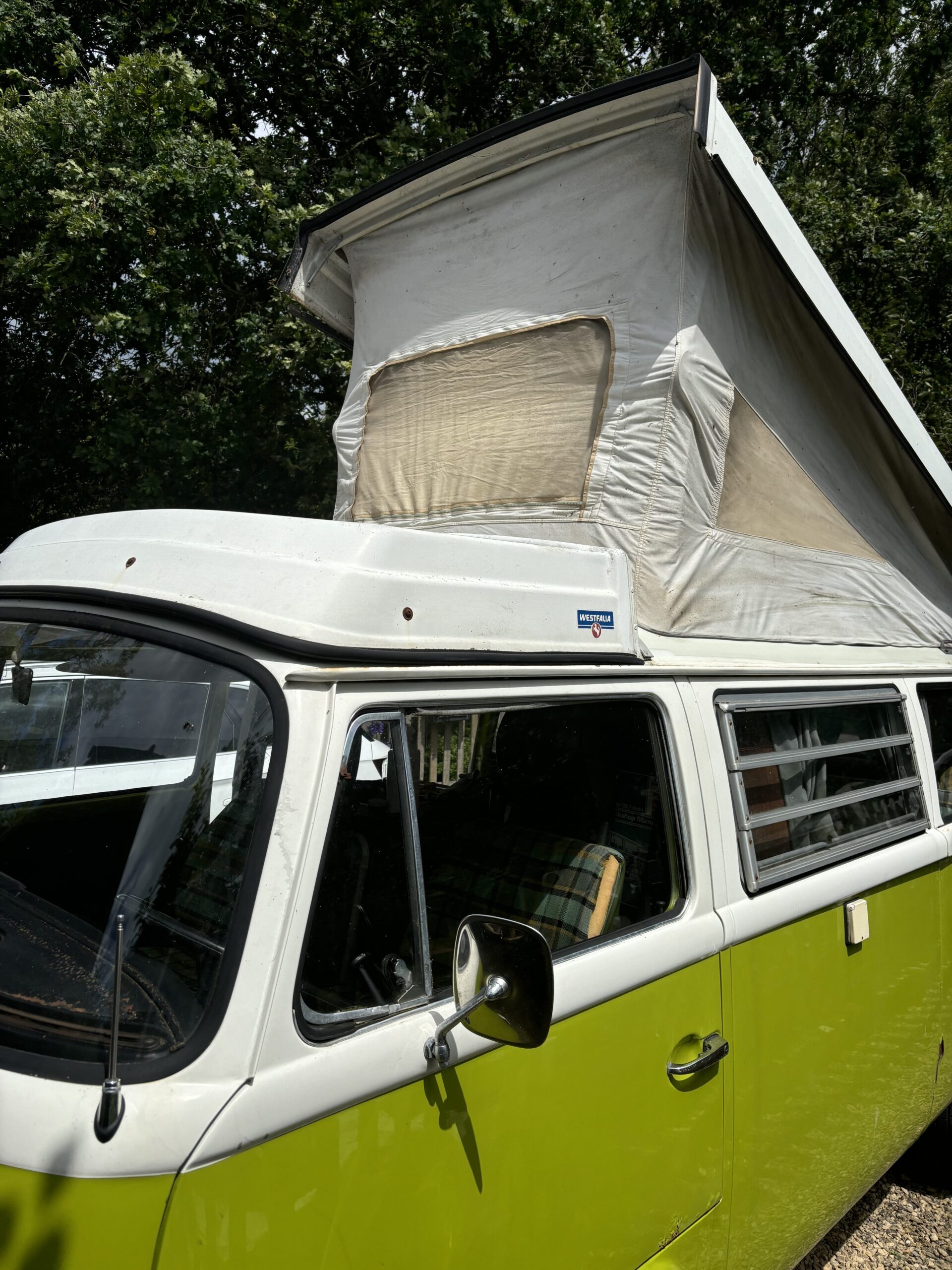 A classic green and white Volkswagen camper van with a raised pop-up roof is parked outdoors. The pop-up roof has a beige canvas with a window, allowing for additional headroom inside. The side windows are slightly open, and the surrounding area features lush green trees.