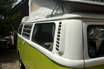 A lime green vintage Westfalia Volkswagen camper van is parked outside on a gravel driveway next to a wooden house. The pop-up roof is extended. It is a sunny day, with a partly cloudy sky visible above. Trees and greenery surround the area, creating a serene outdoor setting.