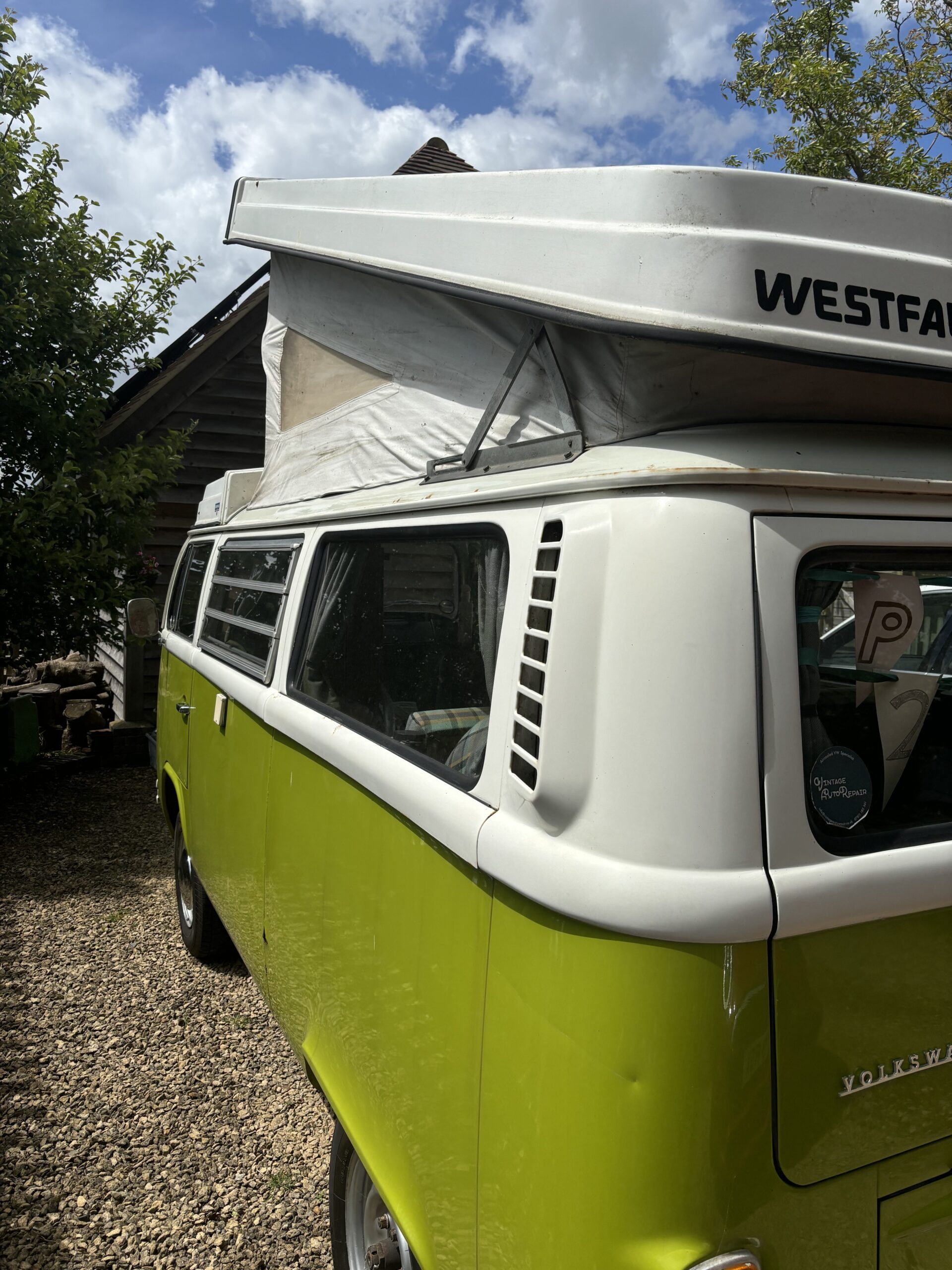 A lime green vintage Westfalia Volkswagen camper van is parked outside on a gravel driveway next to a wooden house. The pop-up roof is extended. It is a sunny day, with a partly cloudy sky visible above. Trees and greenery surround the area, creating a serene outdoor setting.