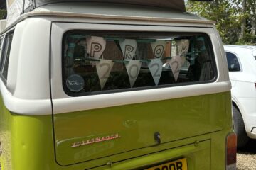 A green and white Volkswagen Westfalia camper van is parked outdoors. The rear window is decorated with triangular flags spelling "PROM 2022". The van has a yellow license plate with the registration "TJT 390R". In the background, there are trees and part of a white car.